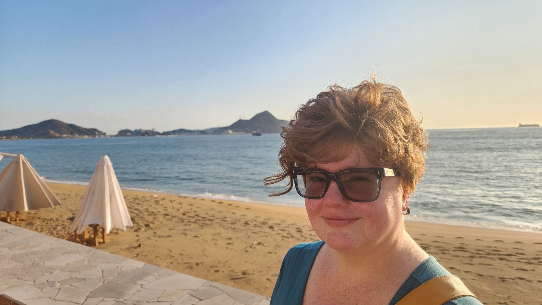 Renee Shupe in sunglasses on a beach at golden hour, the ocean and hills behind her