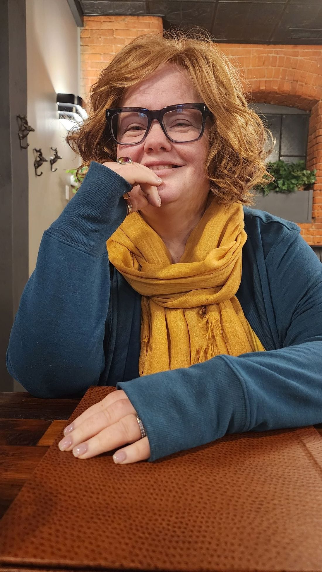 Renee Shupe smiling, chin on her hand, in a mustard scarf in front of a brick wall