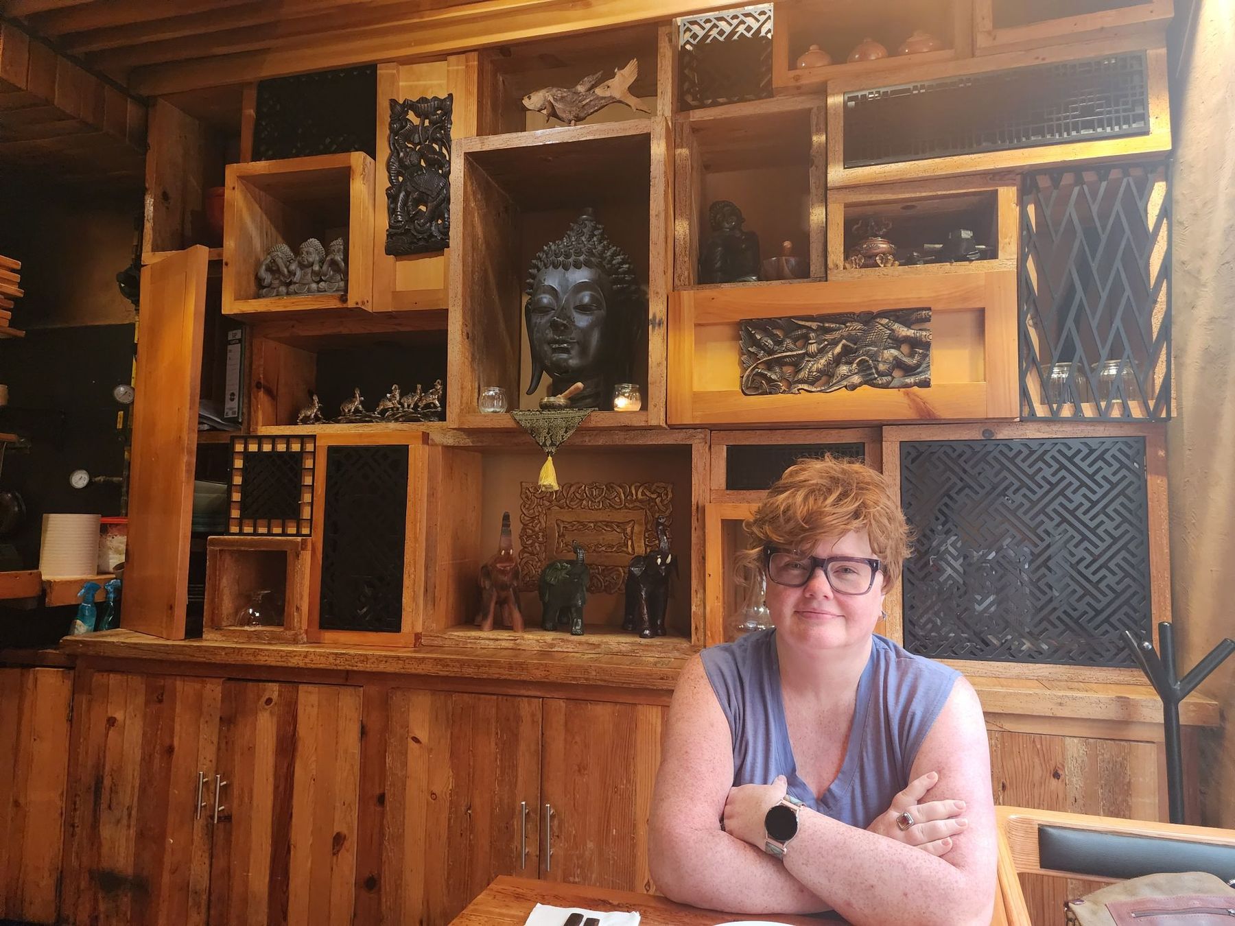 Renee Shupe sitting with arms folded in front of a wall of wooden shelves and carvings