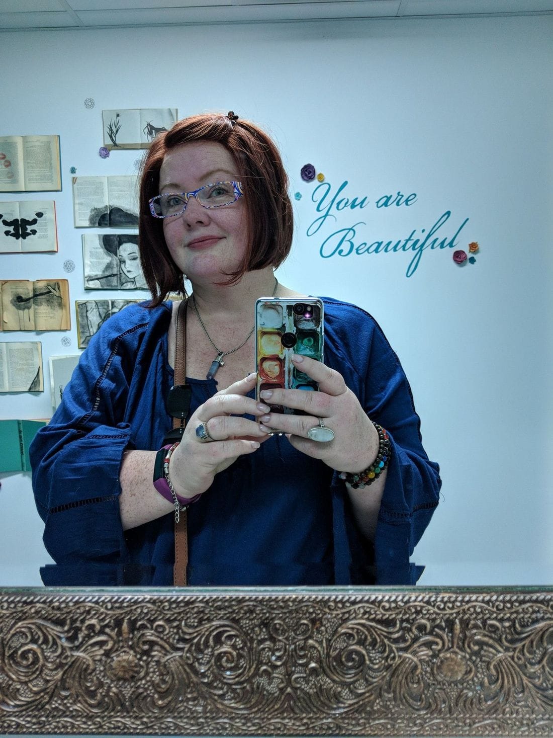 Renee Shupe taking a mirror photo in a blue jacket, with the words You are Beautiful on the wall behind her